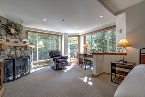 A spacious room with a fireplace, a leather chair, a desk with a lamp, and large windows offering a view of trees. - Woodrun 208 - Ski in Ski out, Common Area Hot Tub & Pool, Gas Fireplace, BBQ (Whistler)