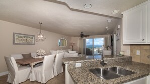 A kitchen with granite countertops, a double sink, and a dining area with white upholstered chairs and a wooden table.