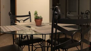 A dining area with a glass table, black chairs, a potted plant, and a framed picture on the wall.