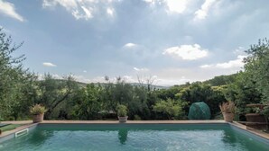 A rectangular swimming pool with clear water, surrounded by lush greenery and a view of distant hills under a partly cloudy sky. - Vacation home Olivo by Interhome (Castellina in Chianti)