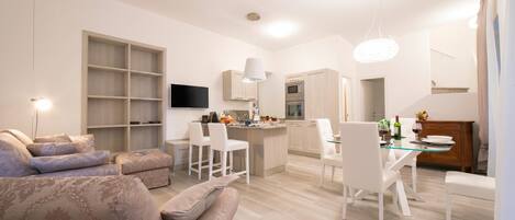 A modern living room with a sofa, a dining area with chairs, a kitchen with a TV, and a staircase.