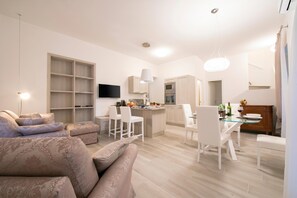 A modern living room with a sofa, a dining area with chairs, a kitchen with a TV, and a staircase.