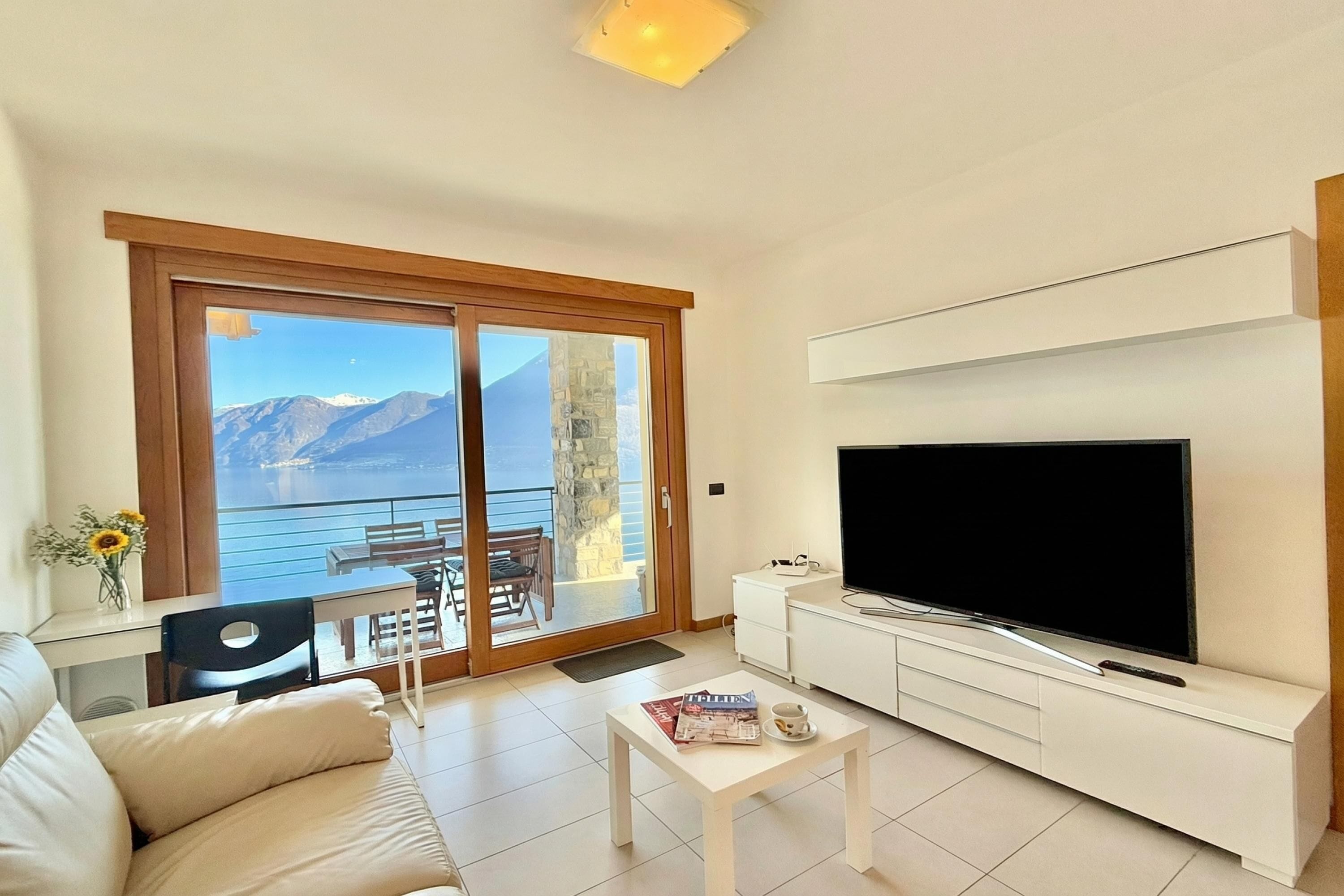 A modern living room with a large TV, a sofa, a coffee table, and a balcony with a mountain view.
