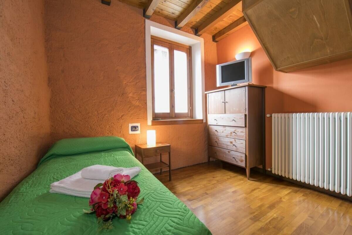Een slaapkamer met een groen bed, een houten dressoir, een televisie op een plank en een raam met zonwering.