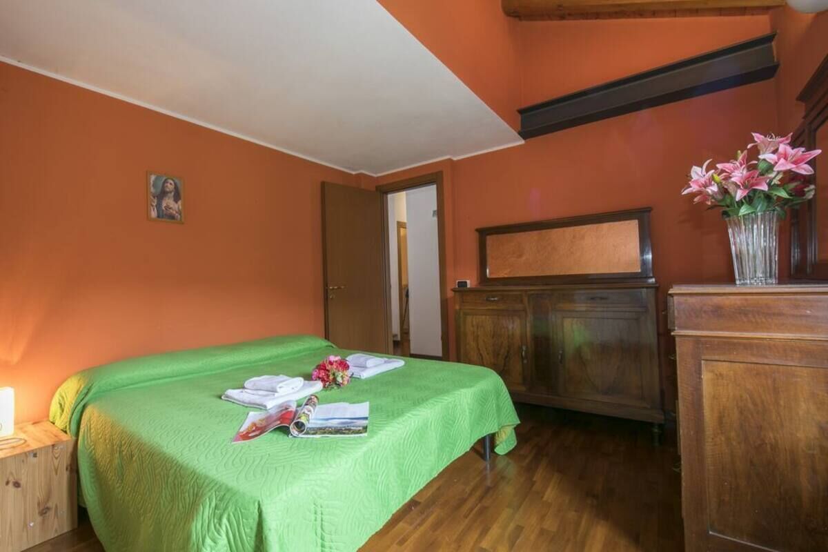 A room with a green bed, wooden furniture, a small painting on the wall, and a vase with flowers on a dresser.