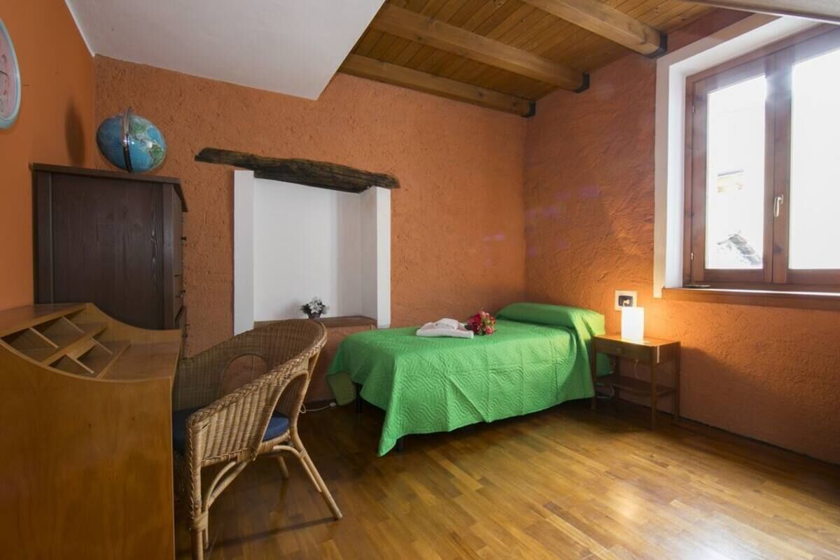 A room with a wooden floor, a bed with green bedding, a wicker chair, a wooden desk, a globe on the wall, and a window with a view.