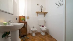 A small bathroom with a sink, bidet, toilet, wooden cabinet, and a shelf with a plant. - Villa Frassi (Pianello del Lario)