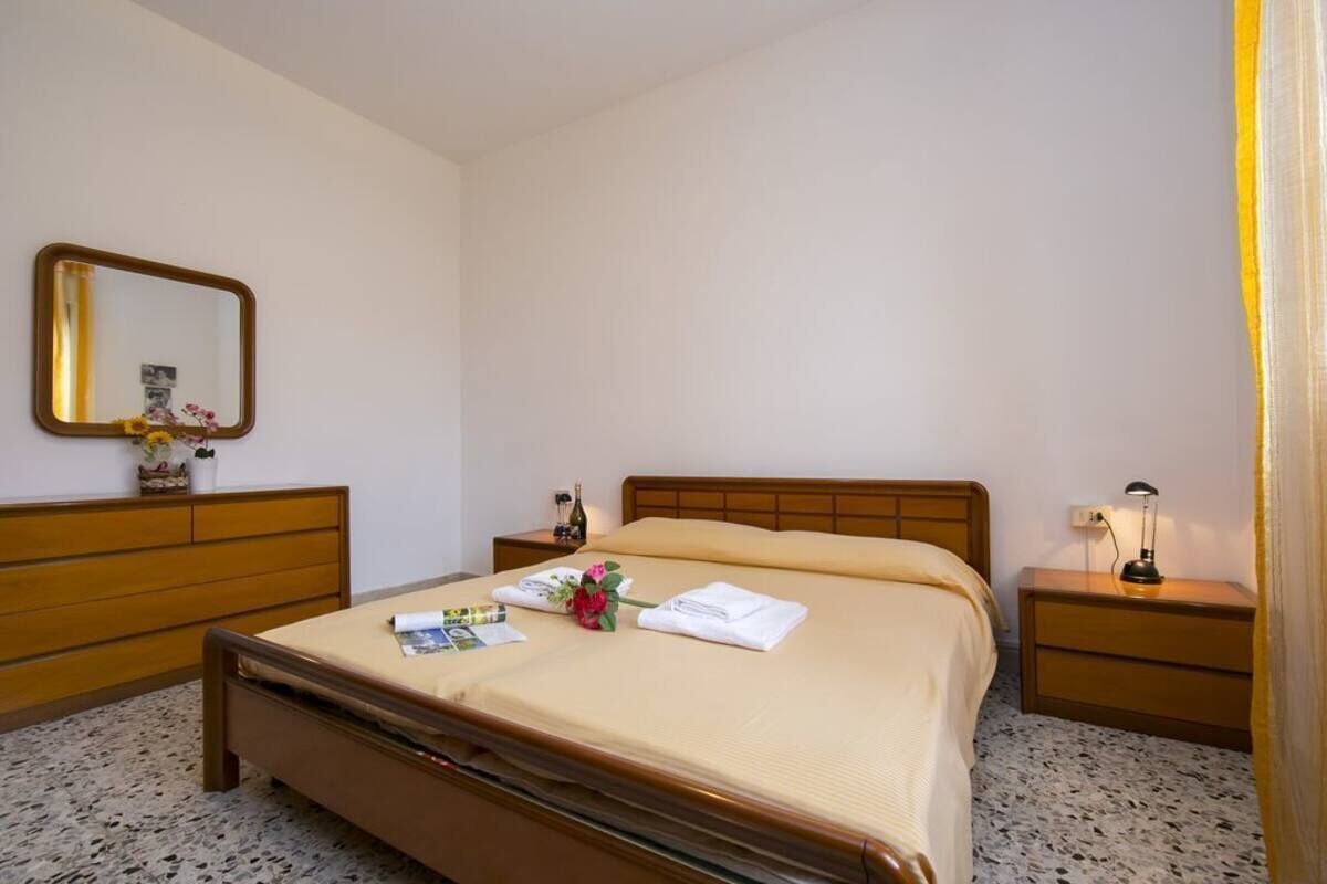 A hotel room with a single bed, a wooden headboard, a nightstand with a lamp, a mirror, and a small table with a bottle and flowers.