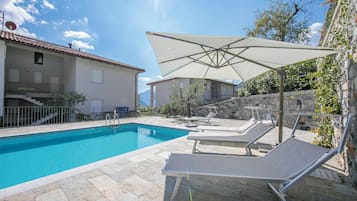 A swimming pool with lounge chairs and a large umbrella, surrounded by stone walls and a building with a tiled roof.