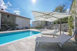 Une piscine dotée de chaises longues et d’un grand parasol, entourée de murs en pierre et d’un bâtiment avec un toit en tuiles.