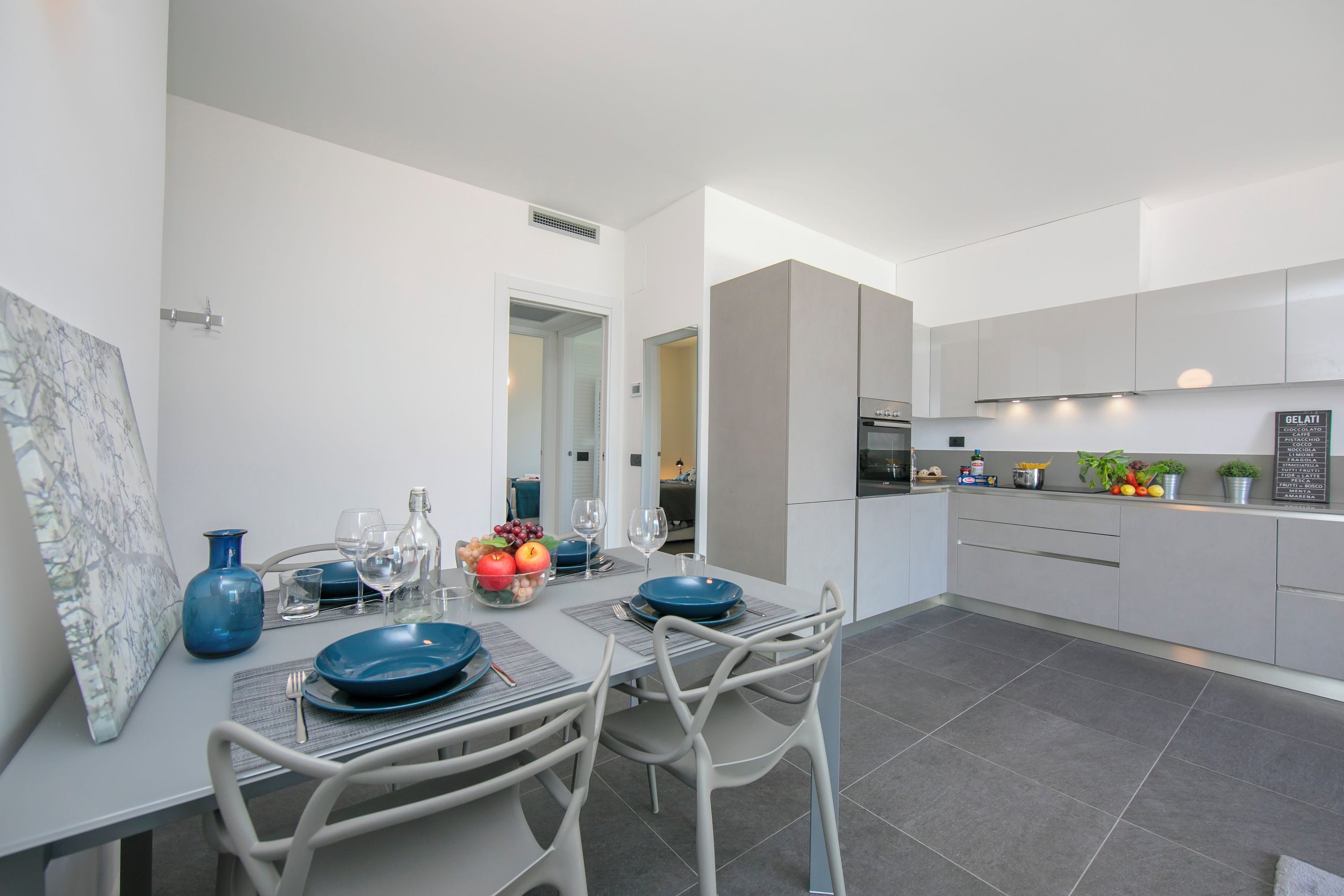 Una cocina moderna con armarios grises, un comedor con una mesa para dos personas y un ventanal grande.