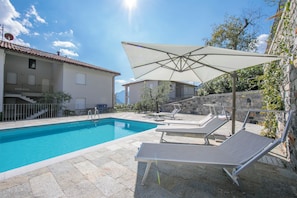 Une piscine dotée de chaises longues et d’un grand parasol, entourée de murs en pierre et d’un bâtiment avec un toit en tuiles.