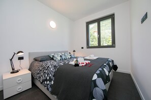 Un dormitorio con cama, mesita de noche, lámpara, ventana y vistas a zonas verdes.