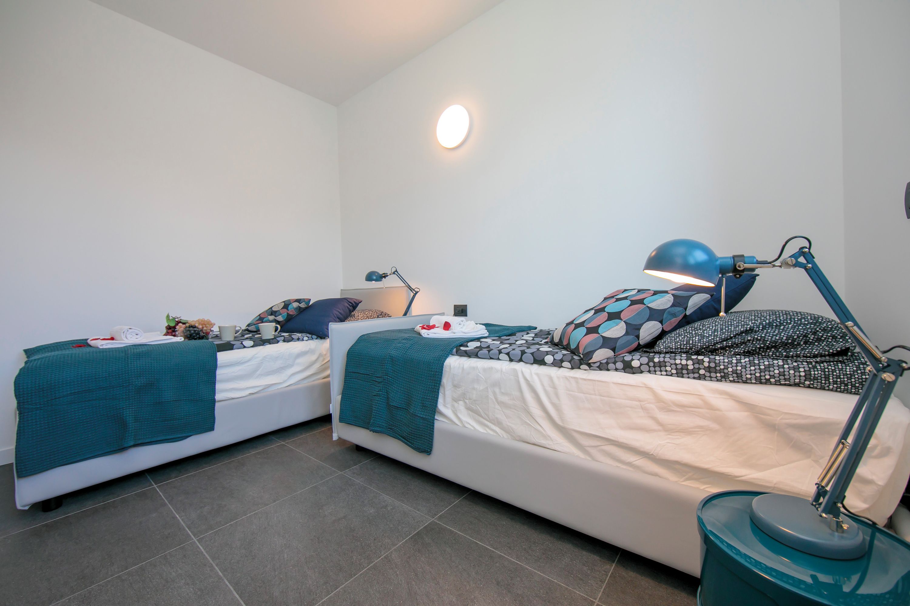 Une petite chambre moderne avec deux lits, chacun recouvert d’une couverture turquoise et agrémenté d’oreillers à motifs. Une lampe de bureau se trouve sur le lit de droite, et une petite table avec une lampe sur le lit de gauche.