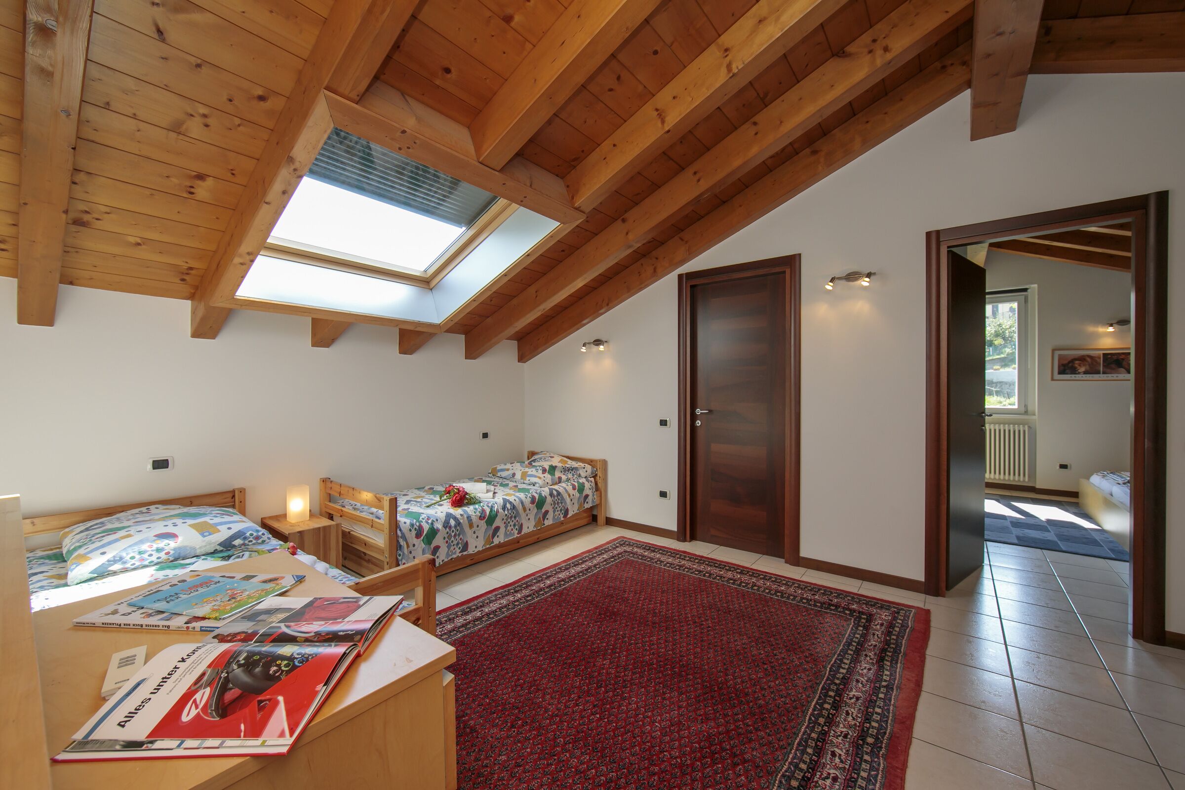 A bedroom with a skylight, two beds, a wooden desk, and a red rug.