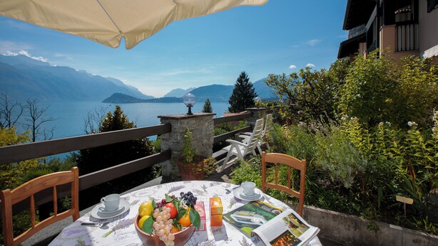 A table with a bowl of fruit, a cup, and a newspaper, overlooking a lake with mountains in the distance. - Sonenga Panoramic Terrace (Menaggio)