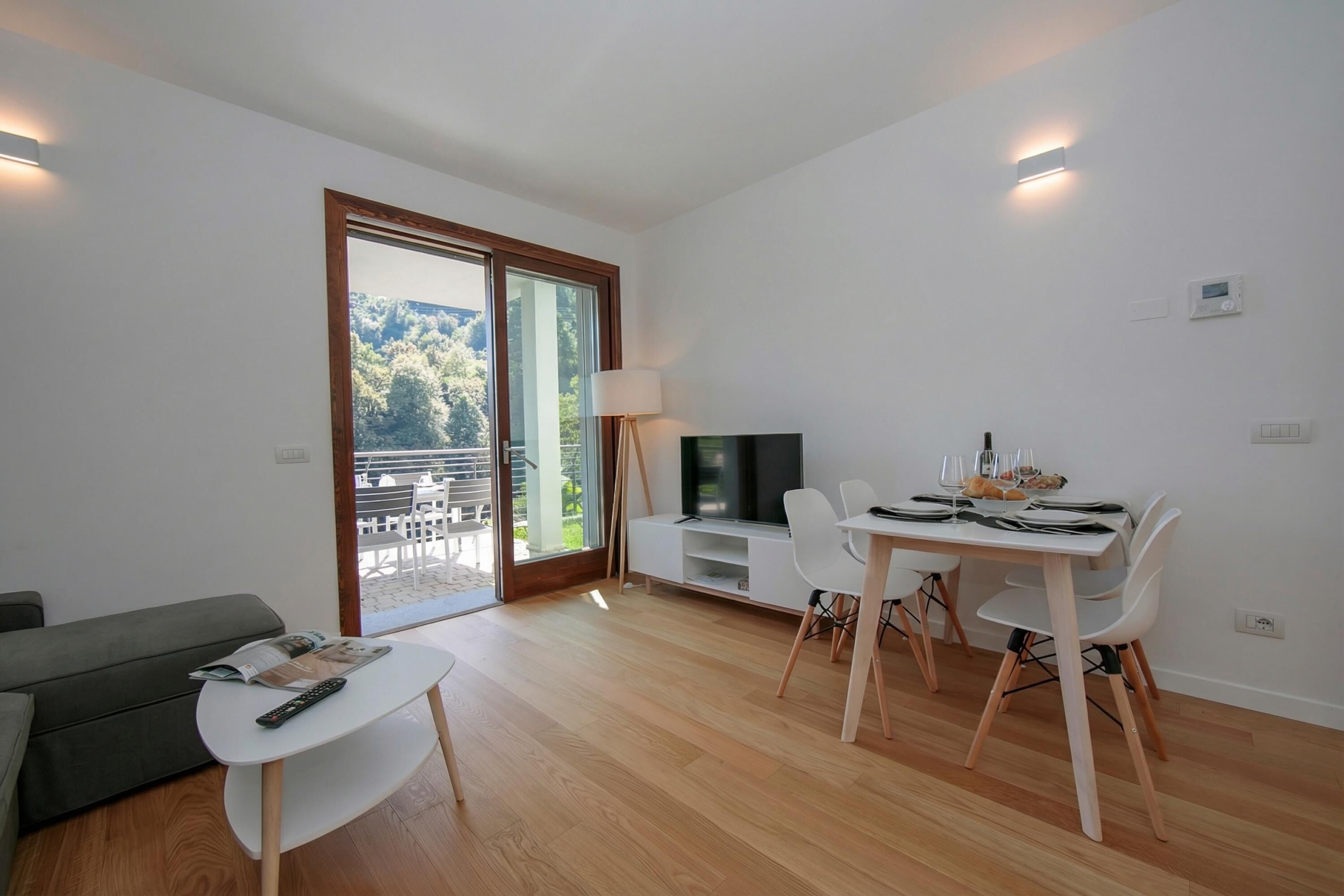 Un soggiorno moderno con un divano, un tavolino, un'area pranzo con sedie, una TV e una porta scorrevole in vetro che dà su uno spazio esterno.