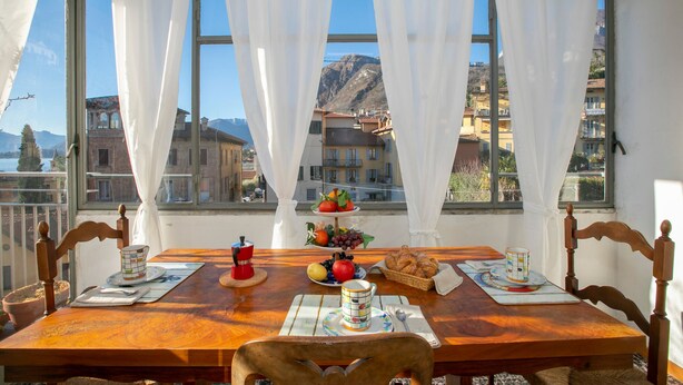 A dining table set with a fruit plate, a coffee pot, and a croissant basket, with a mountain view through large windows. - La Casa del Vitt (Menaggio)