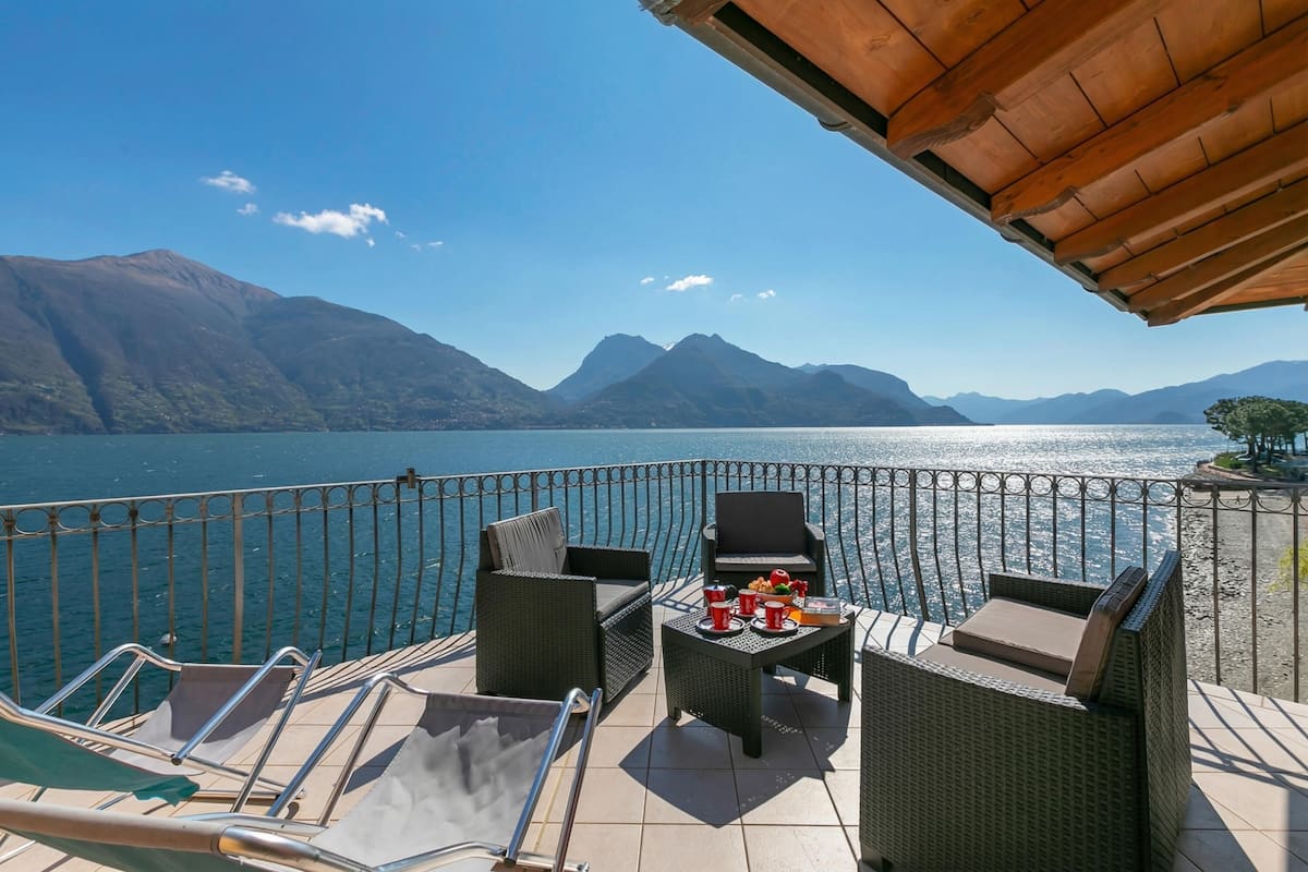 A balcony with wicker furniture, a table with drinks, and a view of a lake and mountains.