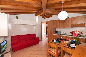 Un ambiente compatto con un divano rosso, un tavolo da pranzo con sedie, un angolo cottura con mobili in legno e un televisore su un supporto.