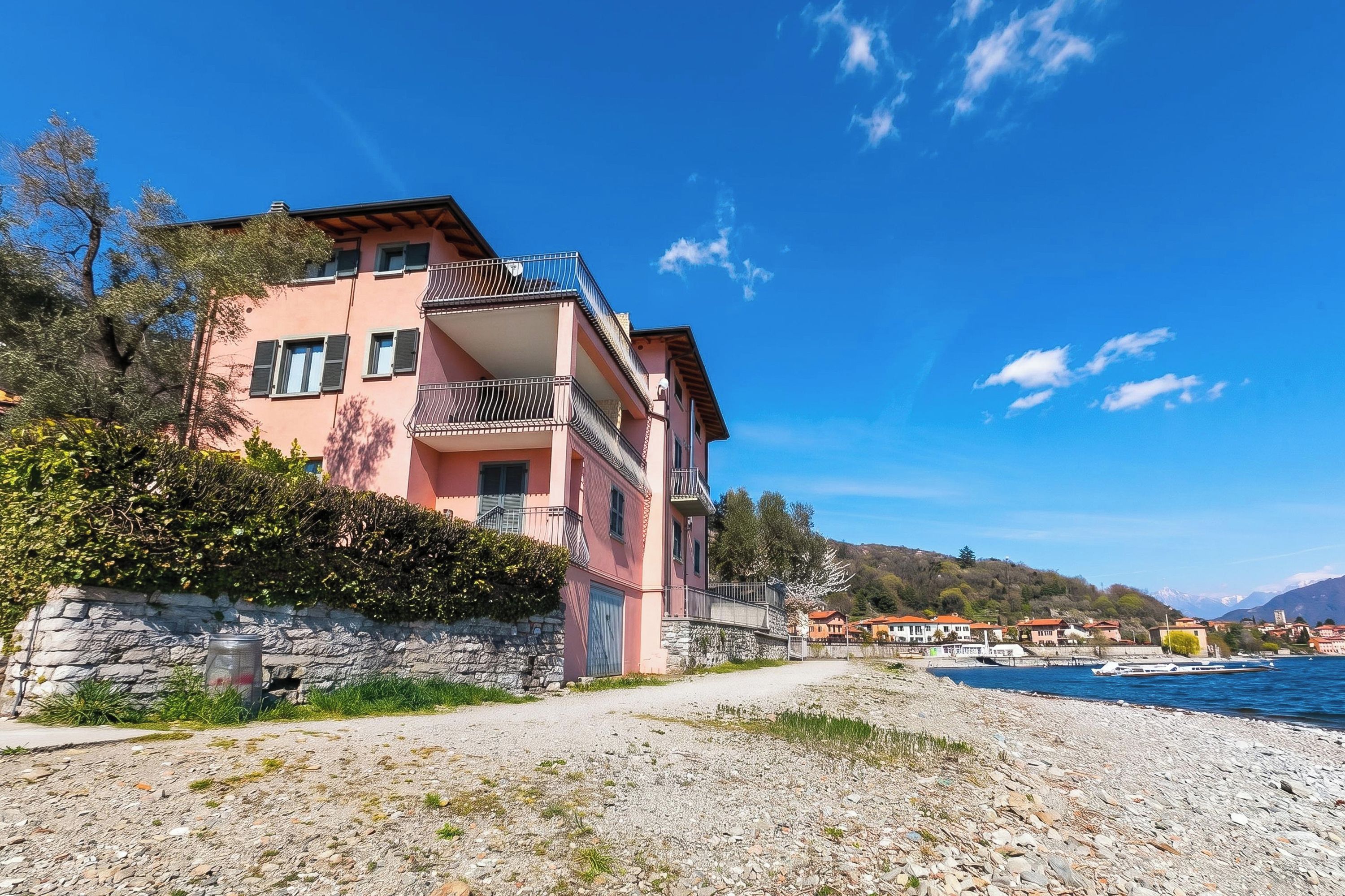 Un edificio rosa a più piani con balconi, un muro in pietra e una spiaggia di ciottoli in primo piano.