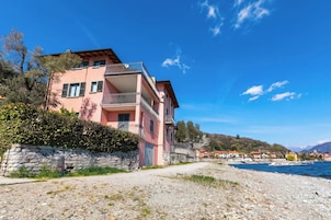 Um edifício cor-de-rosa de vários andares com varandas, uma parede de pedra e uma praia de seixos em primeiro plano.