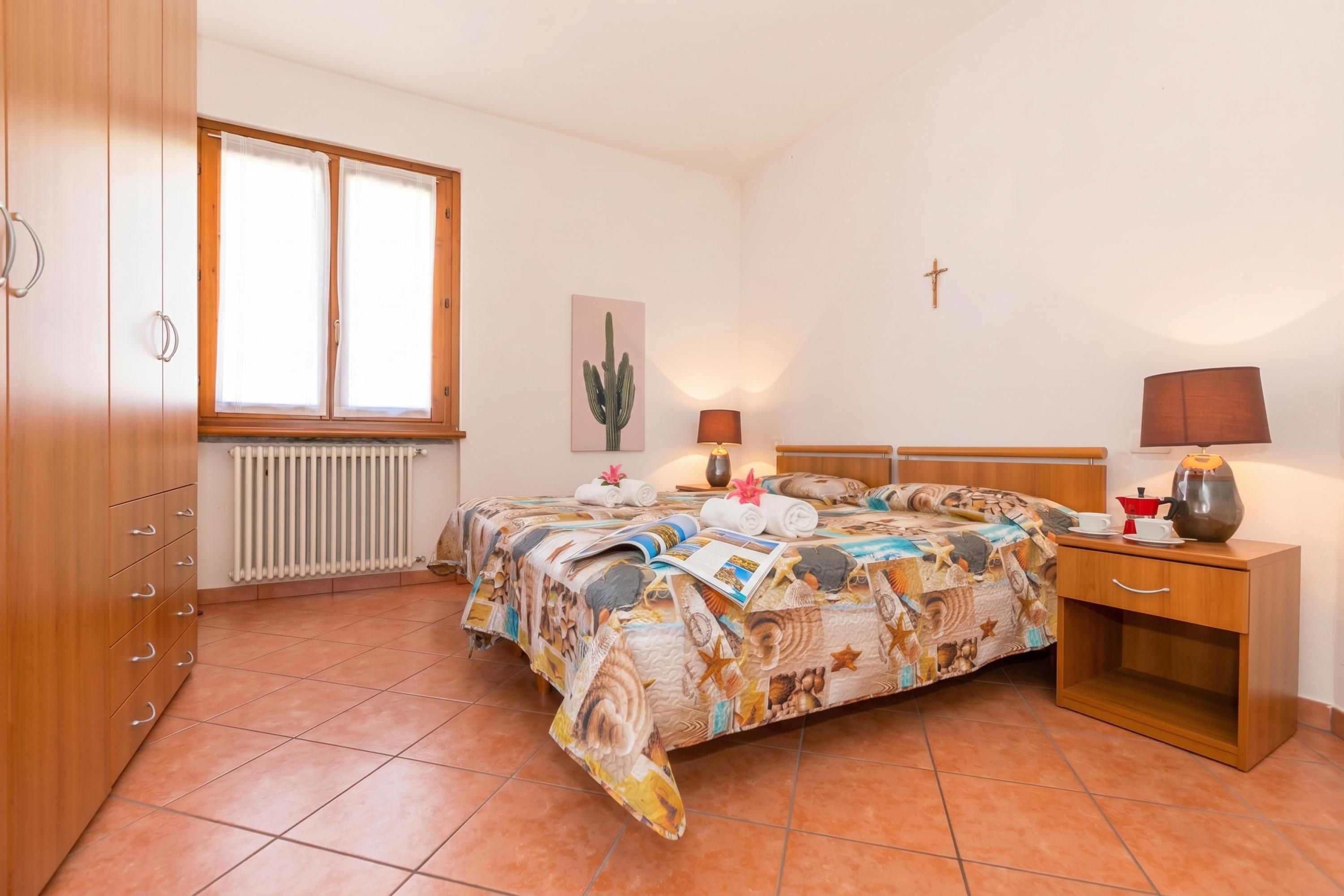Een slaapkamer met een bed, nachtkastjes, een radiator, een raam en een kruis aan de muur.