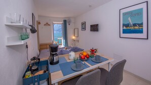 Een kleine kamer met een bed, een eettafel gedekt met borden, kopjes en een waterkoker, een koffiezetapparaat en een klein blauw opbergkastje.