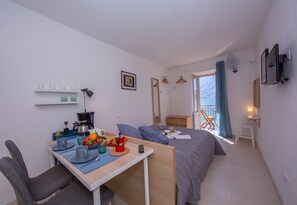 A compact hotel room with a bed, a small desk with a coffee maker, a chair, and a balcony with a view.
