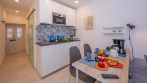 Een compacte keuken met witte kastjes, een magnetron en een blauwe waterkoker. Er is een eethoek met een tafel voor twee personen, een koffiezetapparaat en een afstandsbediening.