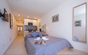 A bedroom with a bed, a large mirror, and a kitchenette in the background.