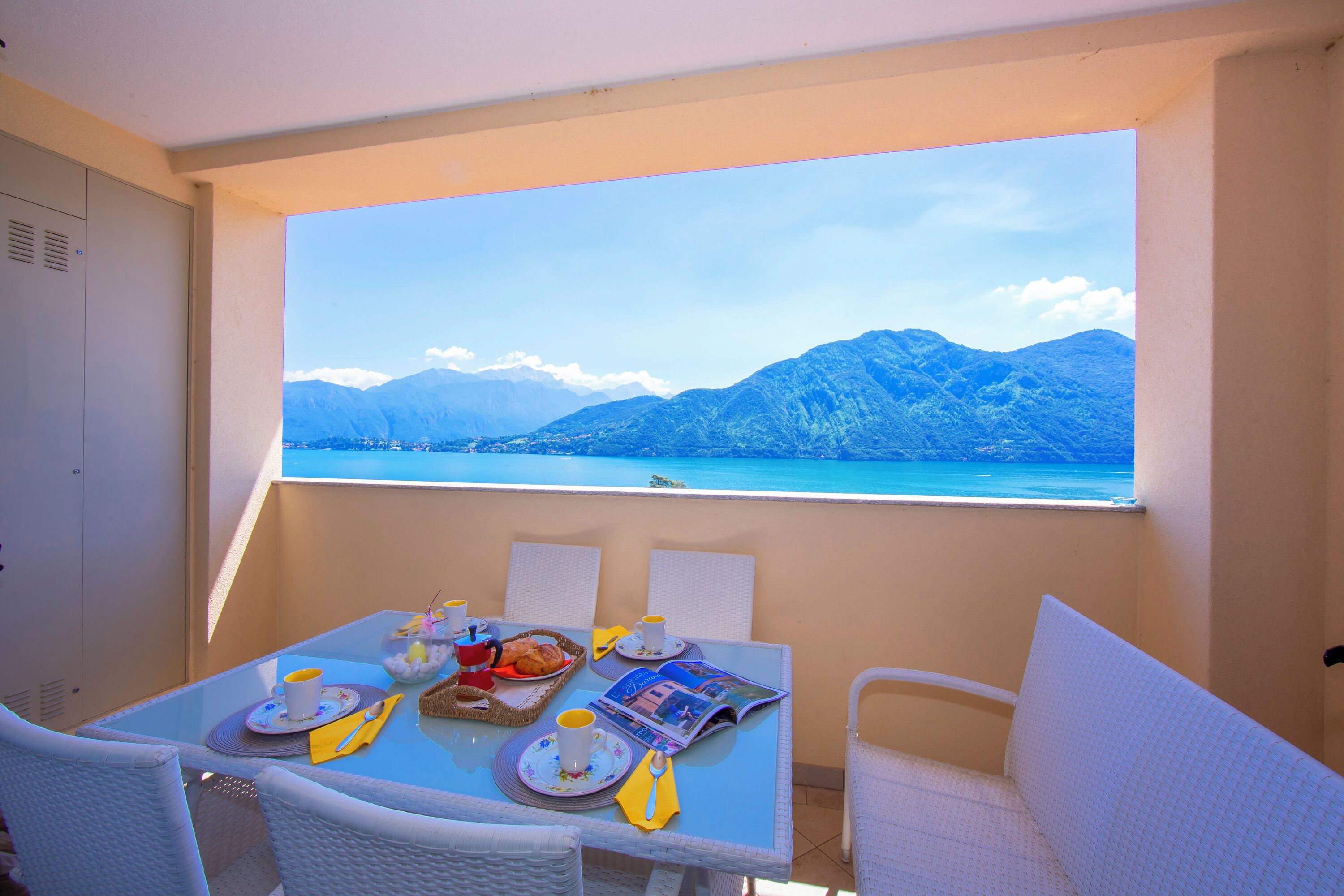 A dining area with a table set for two, a window with a mountain view, and a white wicker chair.