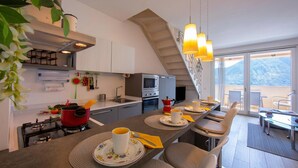 A modern kitchen with a stove, oven, and red pot, a dining area with chairs and a table, and a view of mountains through large windows.