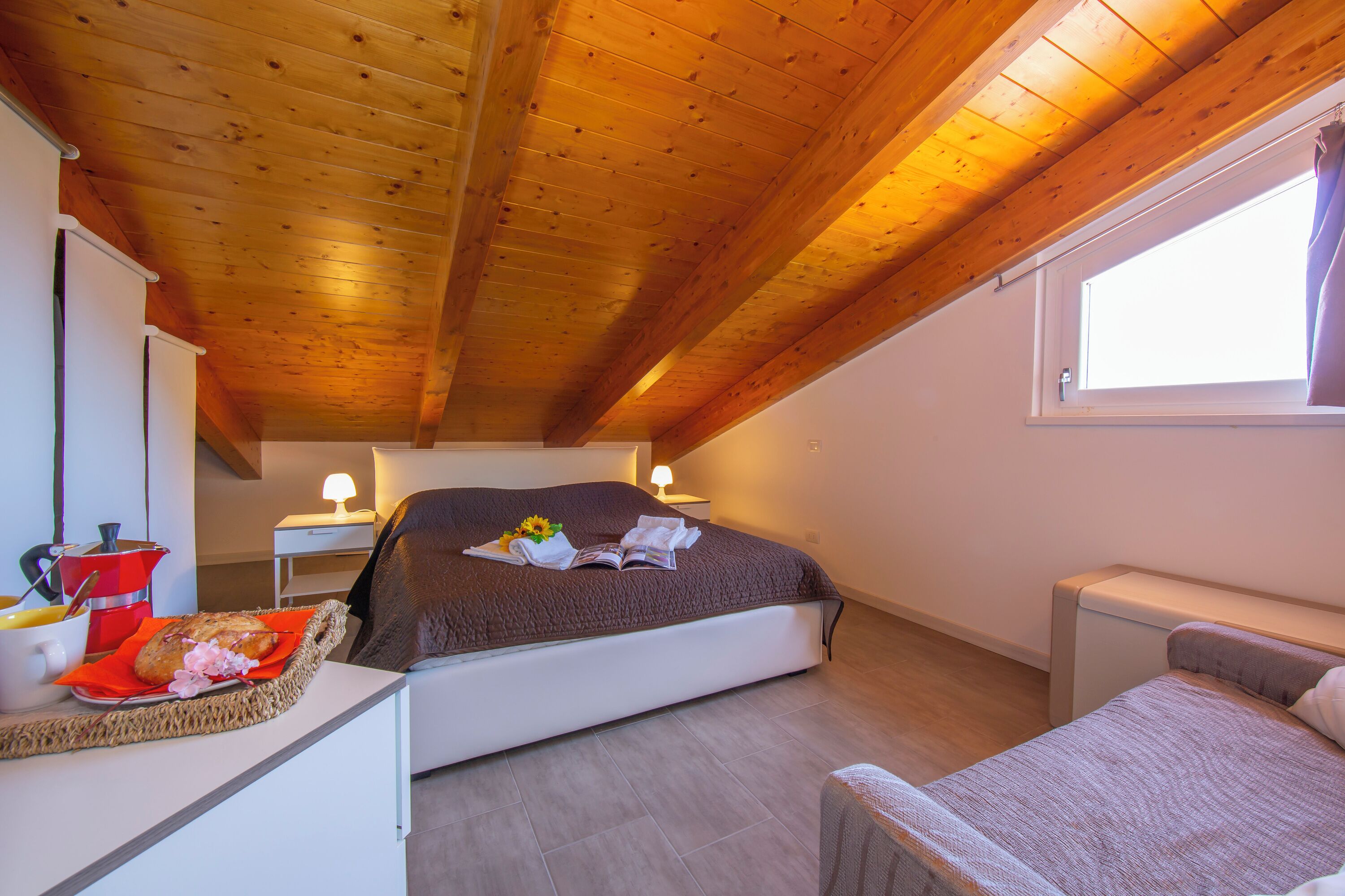 A bedroom with a wooden ceiling, a bed with a brown blanket, a bedside table with a lamp, and a window with curtains.