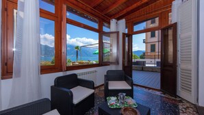 A room with wicker furniture, a table with a tray, and large windows offering a view of mountains and a body of water. - Casa Mira Al Lago (Menaggio)