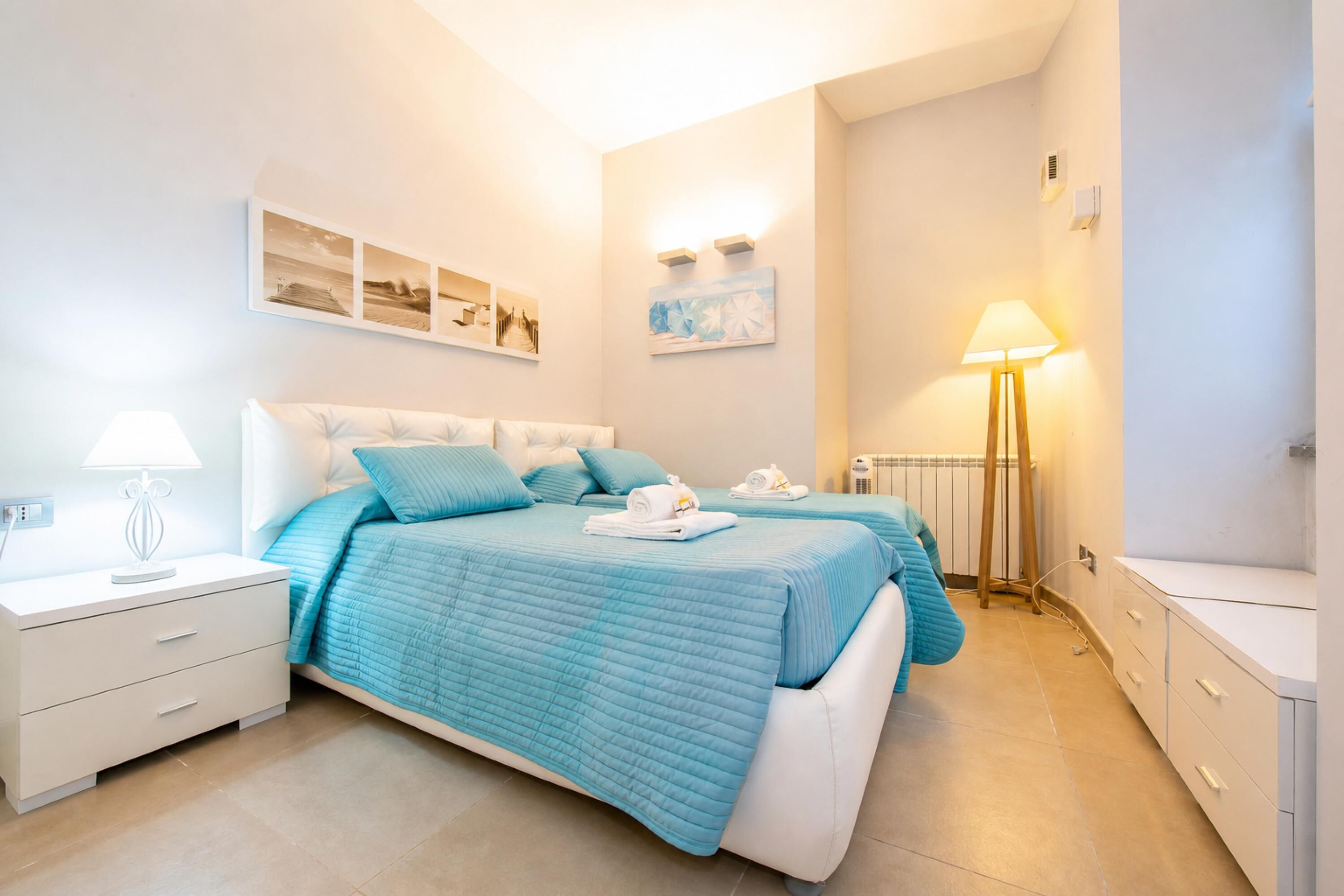 A bedroom with two beds, a bedside table with a lamp, a wall-mounted artwork, and a radiator.