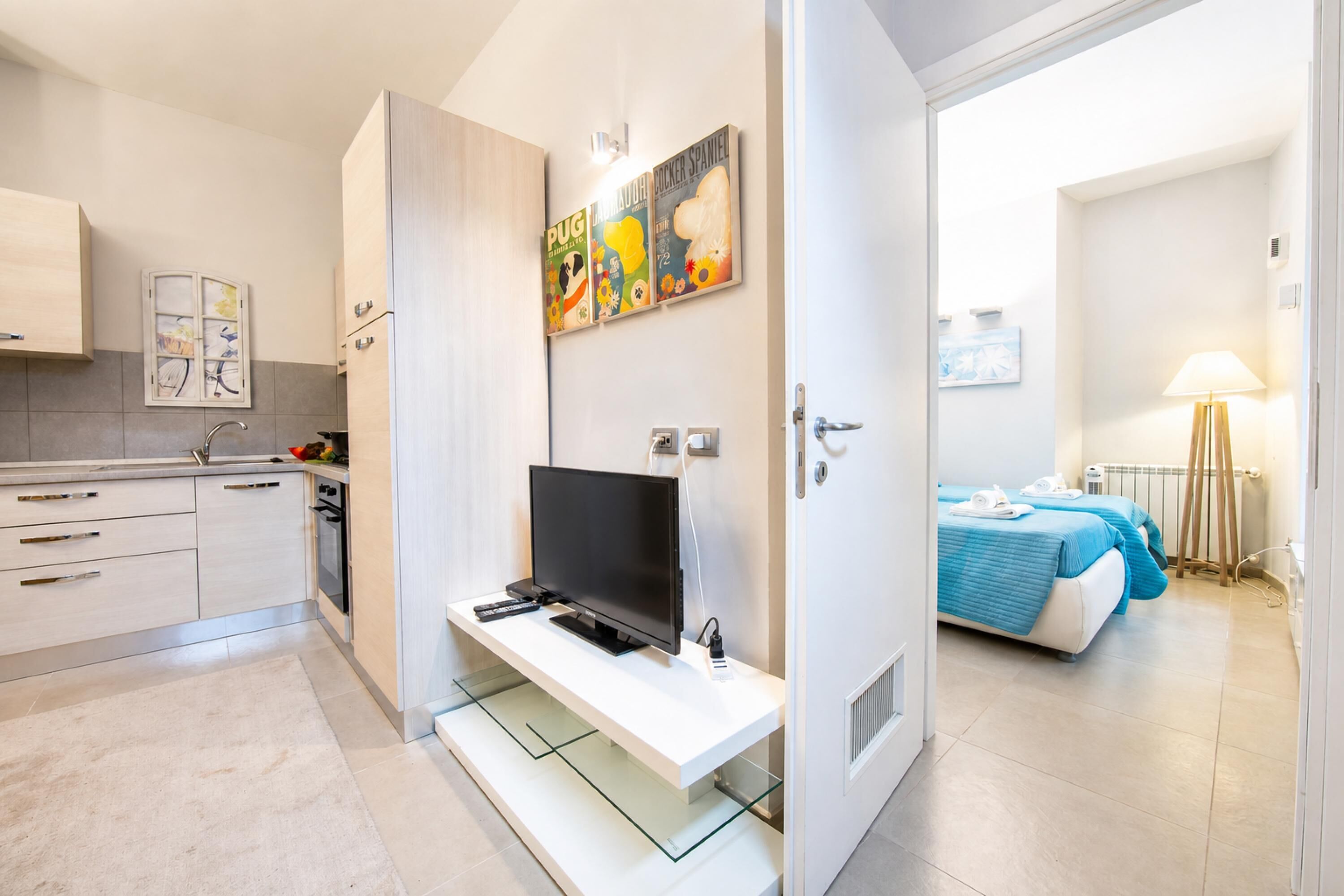 A modern kitchen with a flat-screen TV, a dining area with a table and chairs, and a bedroom with a bed and a bedside table.