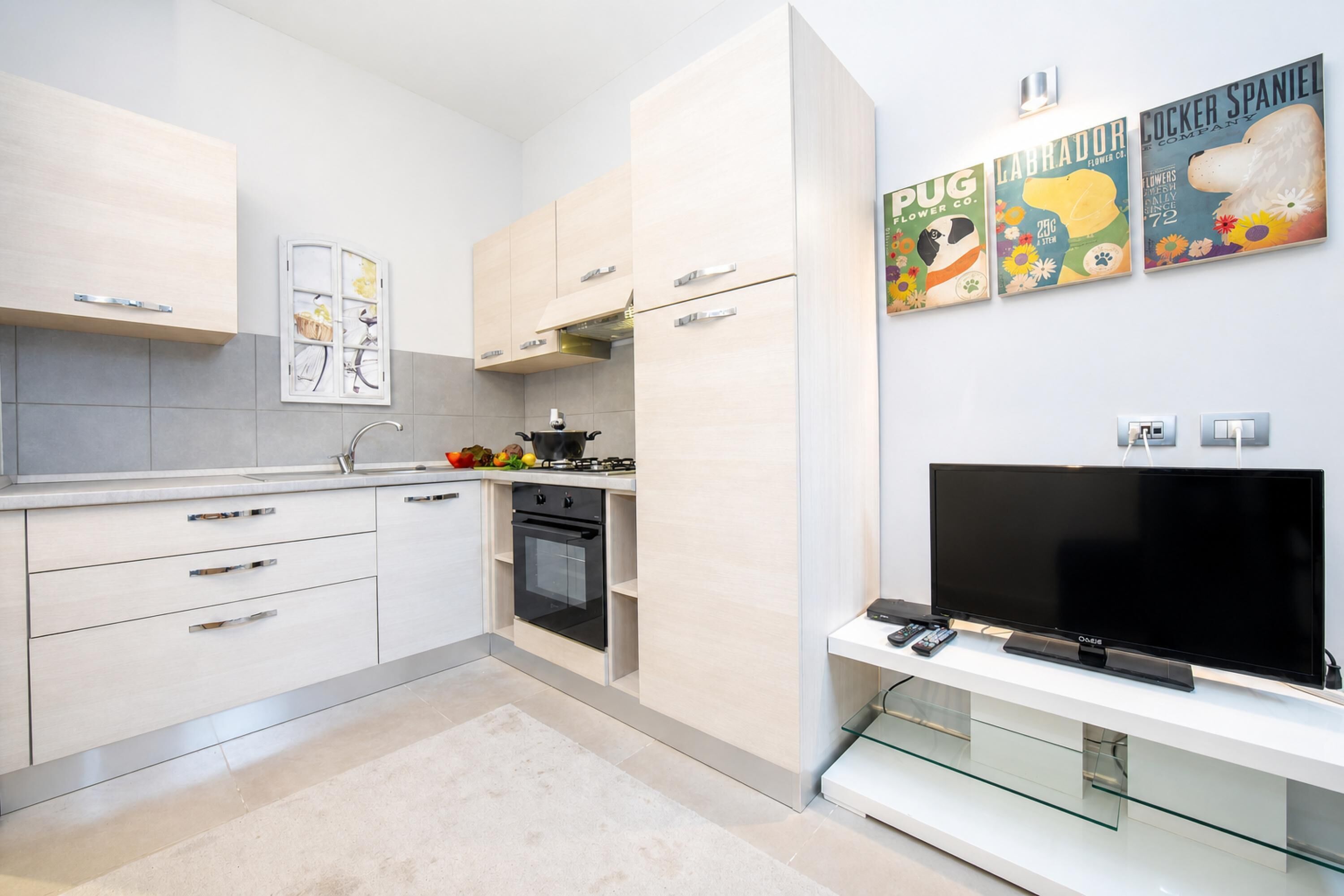 A modern kitchen with white cabinets, a stove, oven, and a flat-screen TV on a glass shelf.