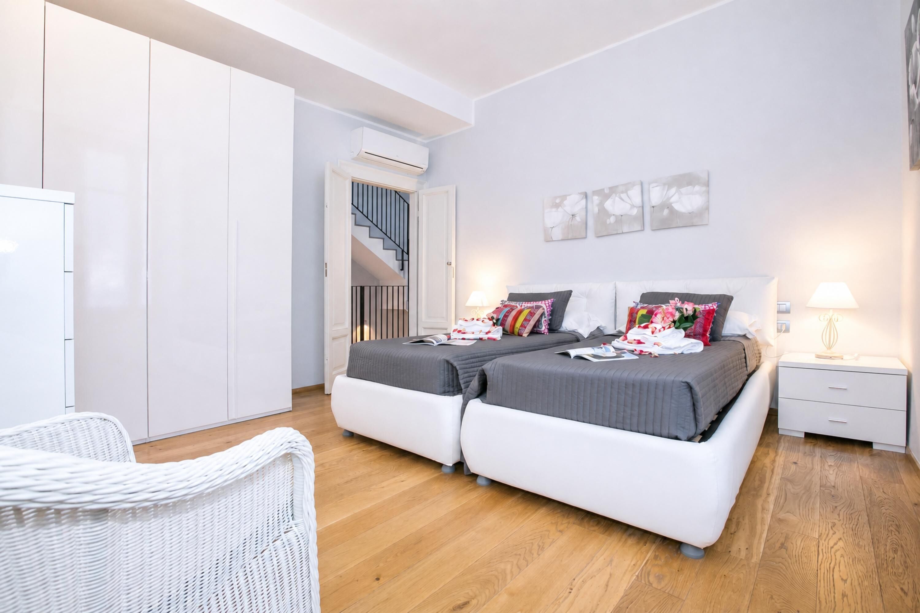 A bedroom with a double bed, a white wardrobe, a white chair, a nightstand, and a staircase leading to an upper floor.