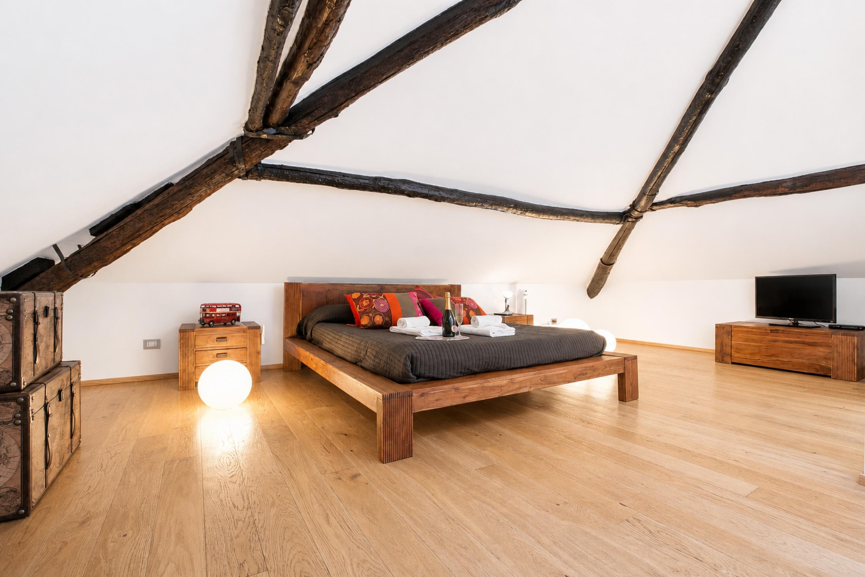 A bedroom with a wooden bed, a bedside table, a nightstand, a television, and a large window.