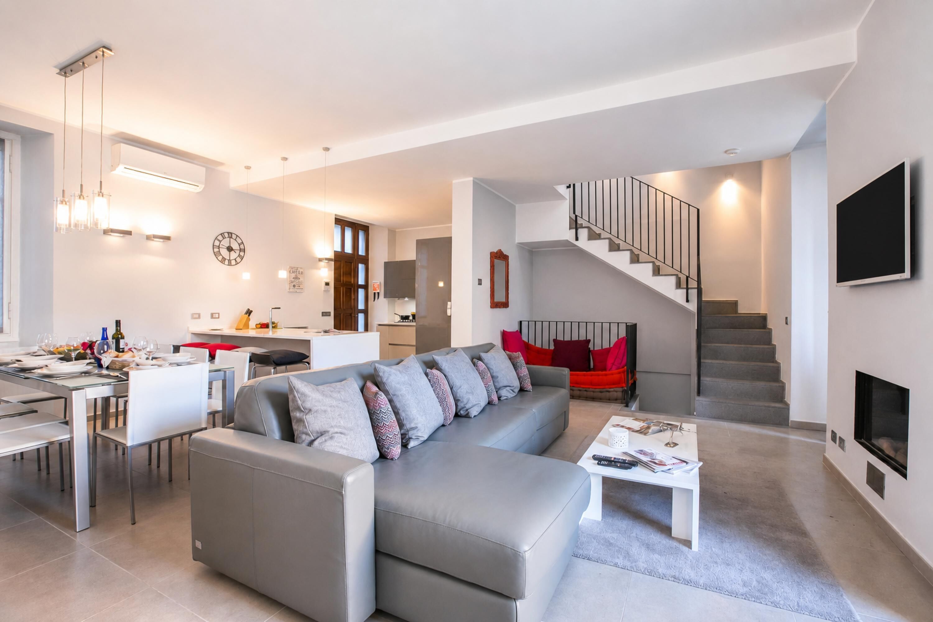 A modern living room with a grey sofa, a dining area, a kitchen, and a staircase.