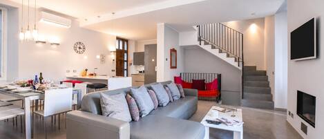 A modern living room with a grey sofa, a dining area, a kitchen, and a staircase.