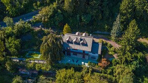 Vista aérea de una casa rodeada de un bosque frondoso, con una piscina visible y un camino de entrada pavimentado.