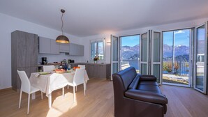 A modern apartment with a dining area, kitchen, and a sofa facing a large sliding glass door with a mountain view.