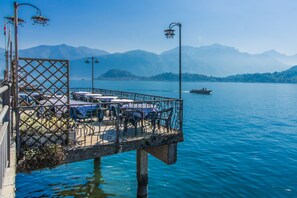 A lakeside terrace with tables and chairs, overlooking a calm lake and distant mountains. - Griante Le Maioliche Apt. 01 (Griante)