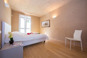 A bedroom with a bed, a chair, a bedside table with a magazine, a potted plant, and a framed picture on the wall. - Griante Le Maioliche Apt. 01 (Griante)