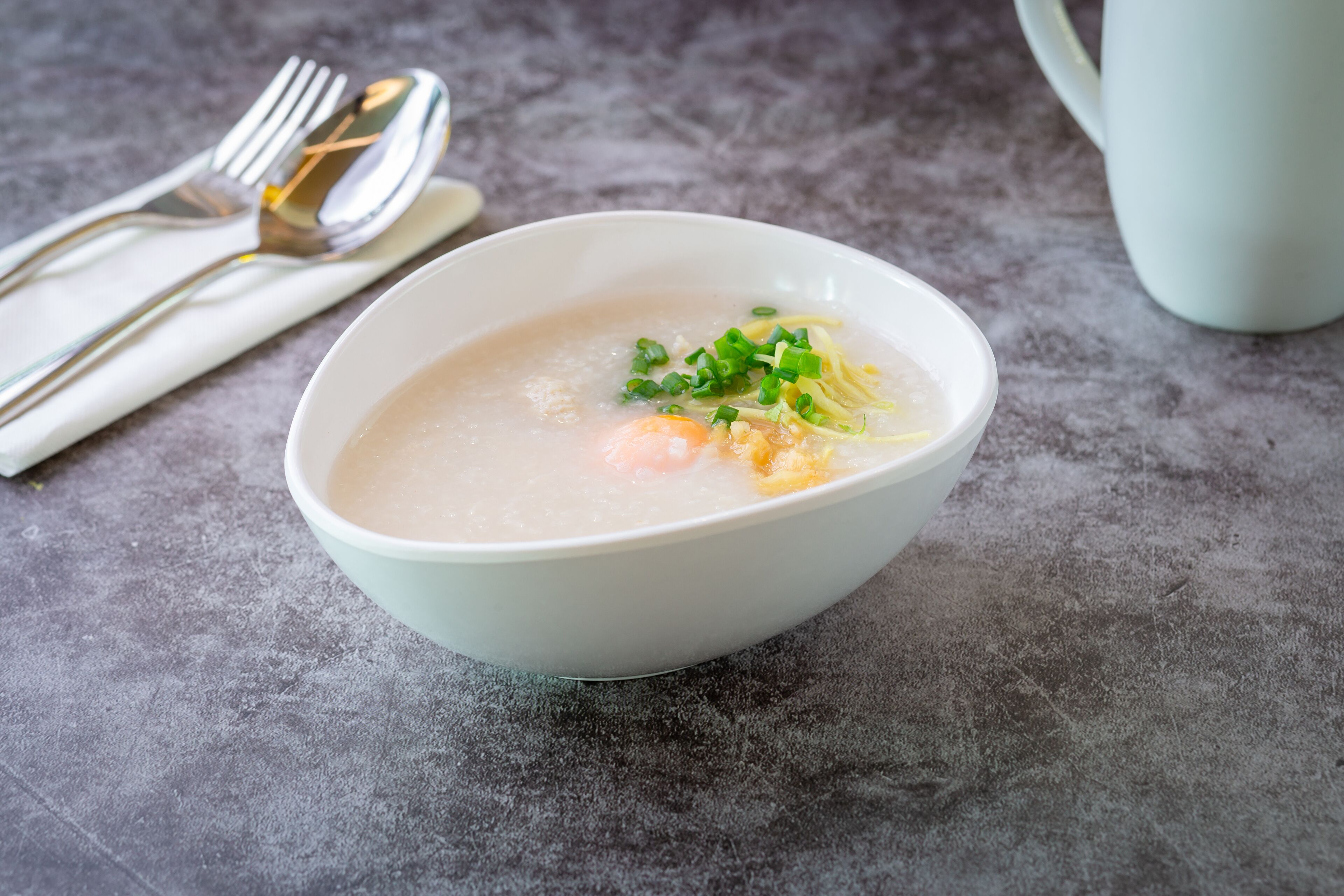 Un tazón de sopa clara con huevo y cebollín, sobre una superficie texturizada, con una taza blanca y utensilios al fondo.