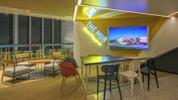 Un espacio de oficina moderno con una pantalla grande mostrando el exterior de un edificio, una zona de estar con diferentes sillas y una pared con un letrero de neón.