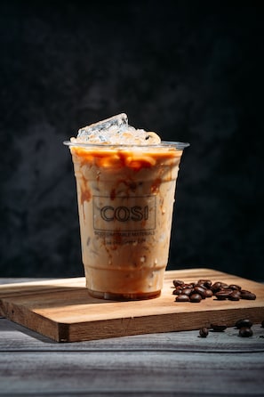 Ein Glas Eiskaffee mit Eiswürfeln auf einer Holzbrettchen mit verstreuten Kaffeebohnen.
