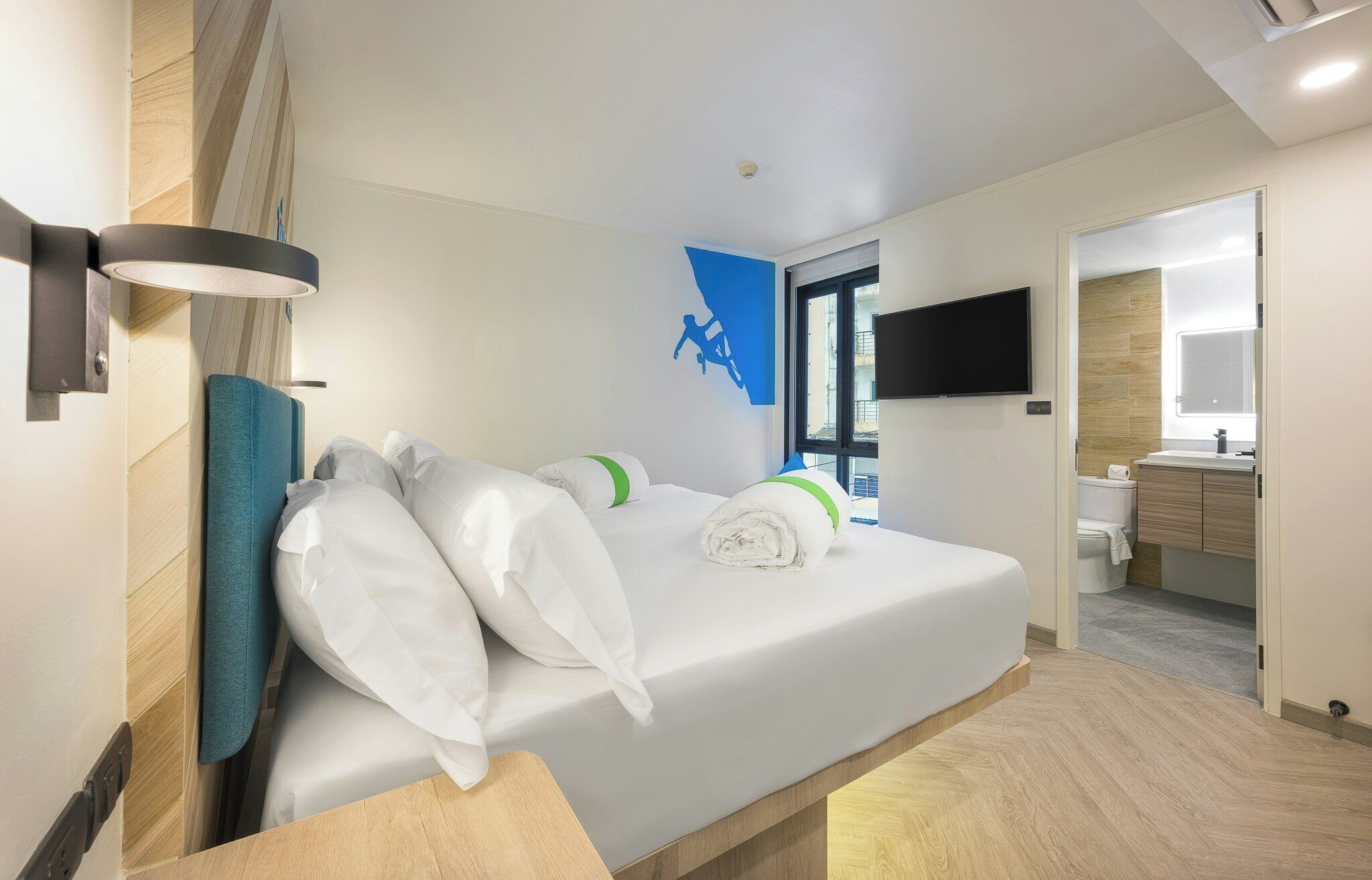 A hotel room with a bed, a flat-screen TV mounted on the wall, and a bathroom visible through an open door.